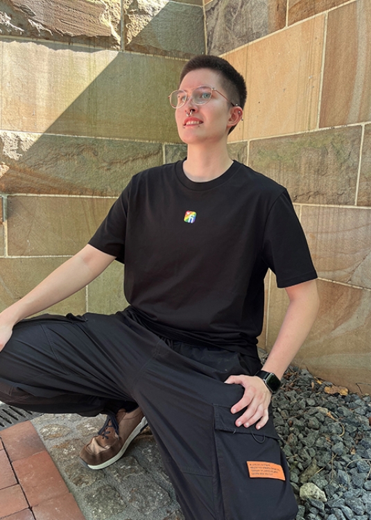 Model vor Steinwand mit schwarzem T-Shirt mit Finanz Informatik Logo in Regenbodenfarben